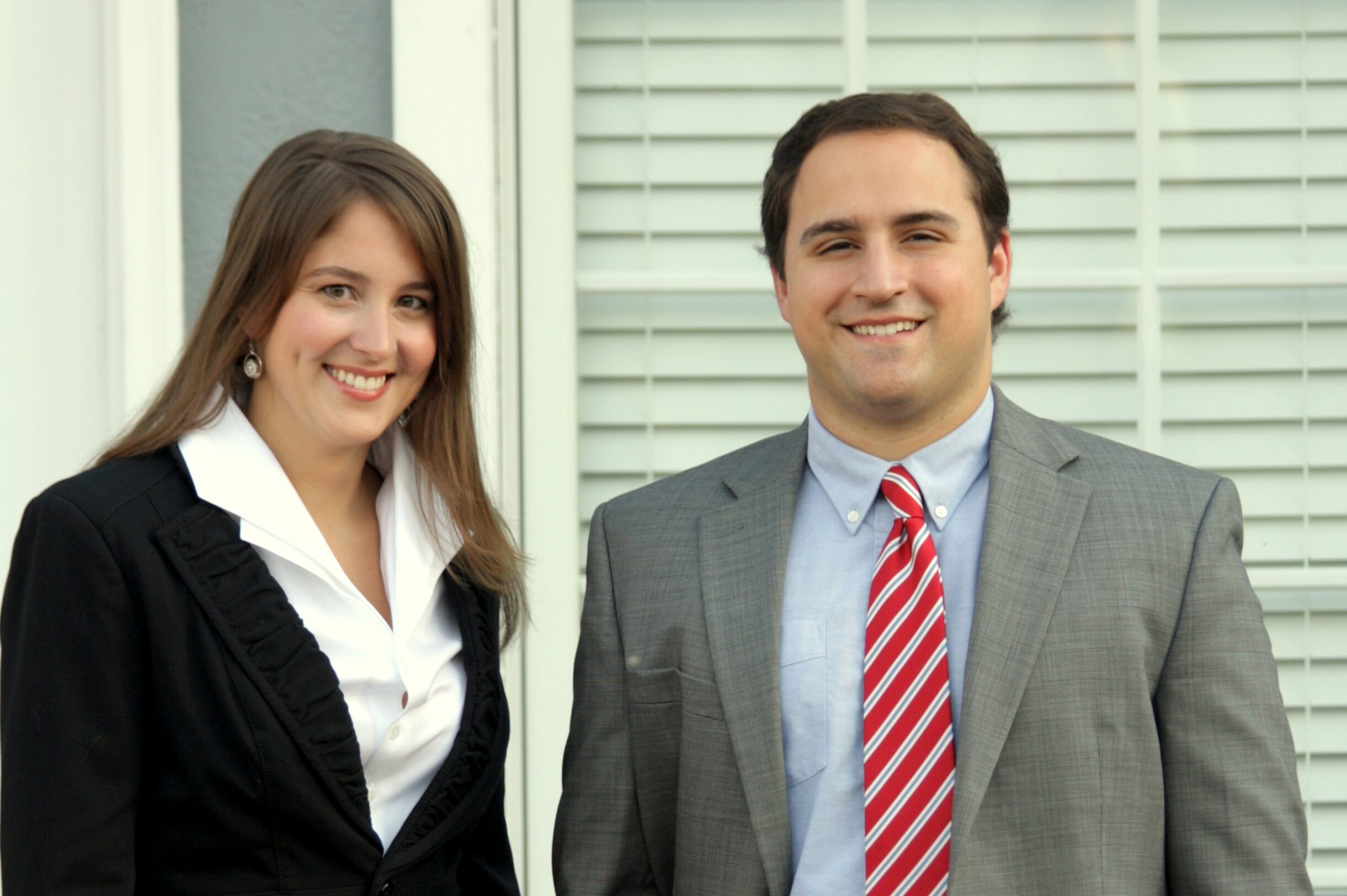 Alyssa and Adam (two attorneys at law with Baxley Maniscalco) are seen posing for a picture. Alyssa is wearing a white blouse with a black top, and Adam is wearing a grey blazer with a red and white tie and light blue shirt.