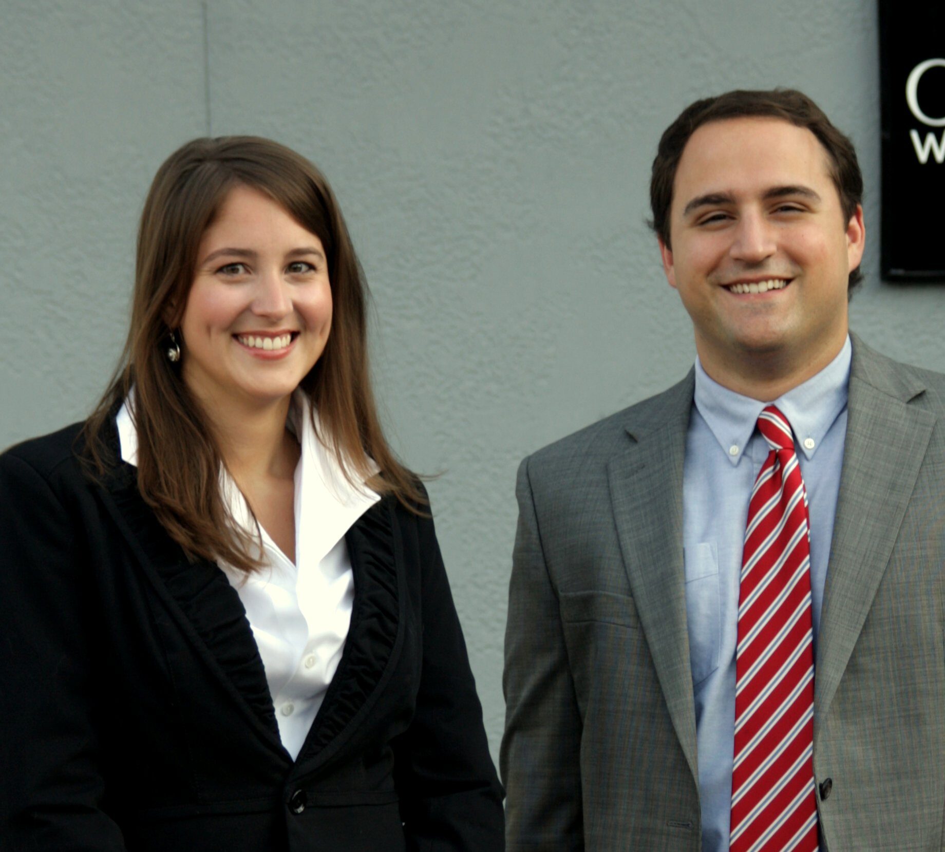 Alyssa & Adam (attorneys at law with Baxley Maniscalco) stand posing for a picture. Alyssa is wearing a white blouse with a black top, and Adam is wearing a grey blazer with a light blue top and a white and red tie.