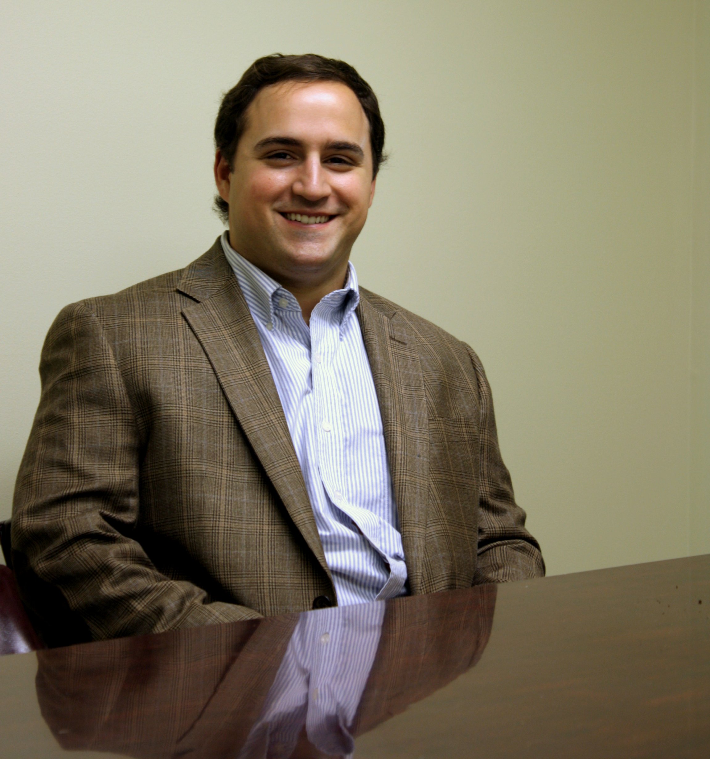 Adam (an attorney at law with Baxley Maniscalco) sits at a table and poses for a picture. He's wearing a brown blazer and a light blue shirt.