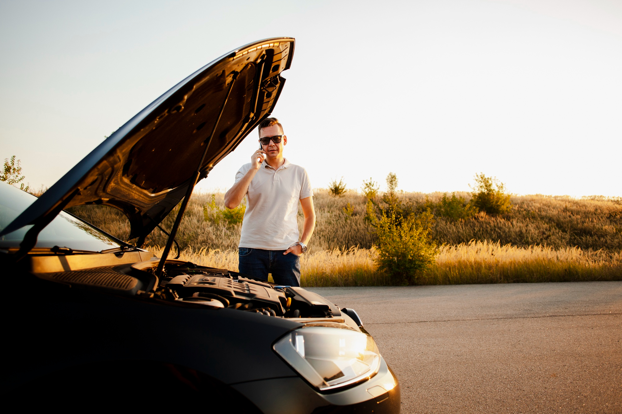 A man stands next to his broken down car on the side of the road. The hood of the car is open.