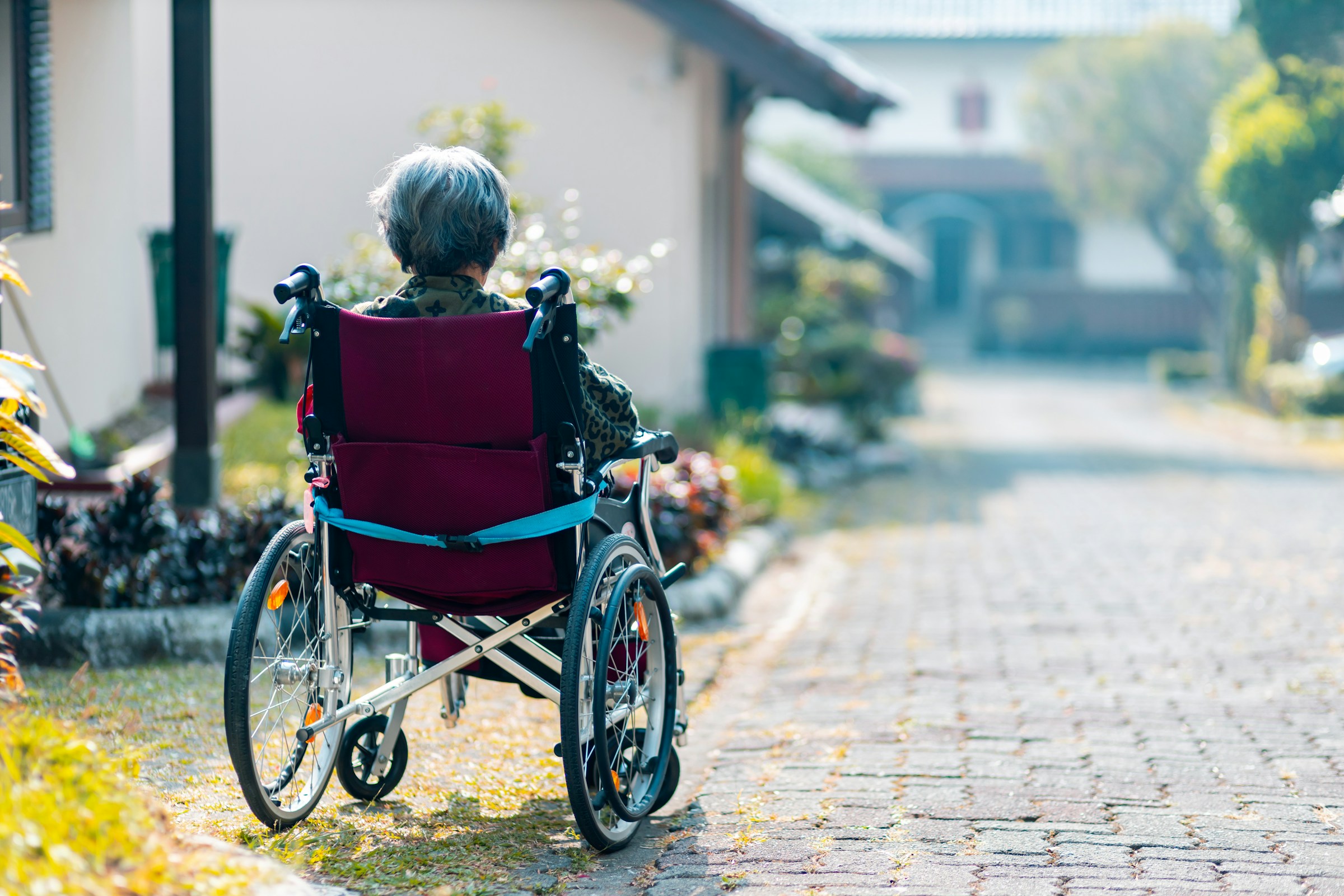 An elderly woman sits in a wheelchair on a cobblestone road. A few houses and structures are seen surrounding her.