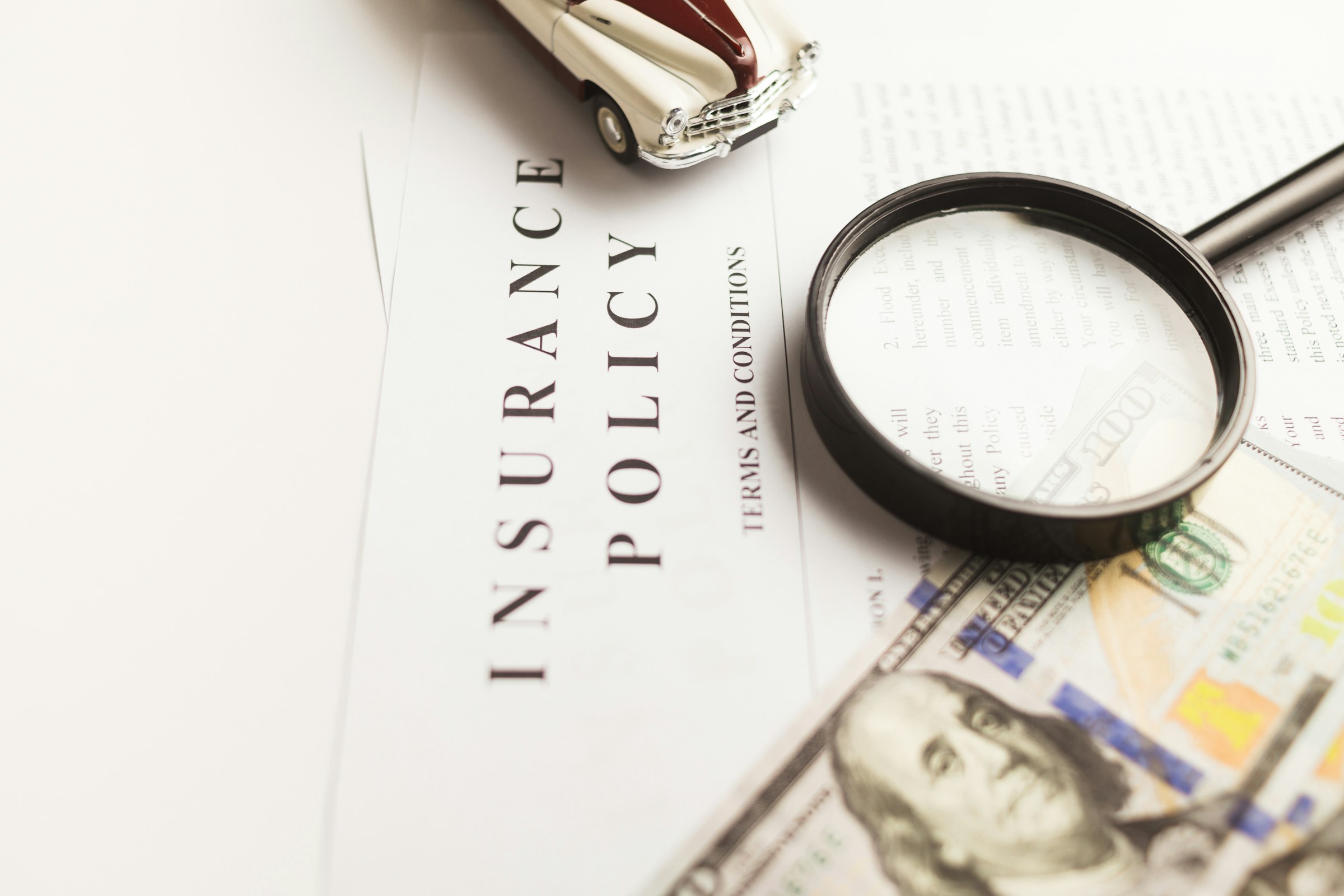 The cover of an insurance policy sits on top of a desk amidst a dollar bill and a magnifying glass.