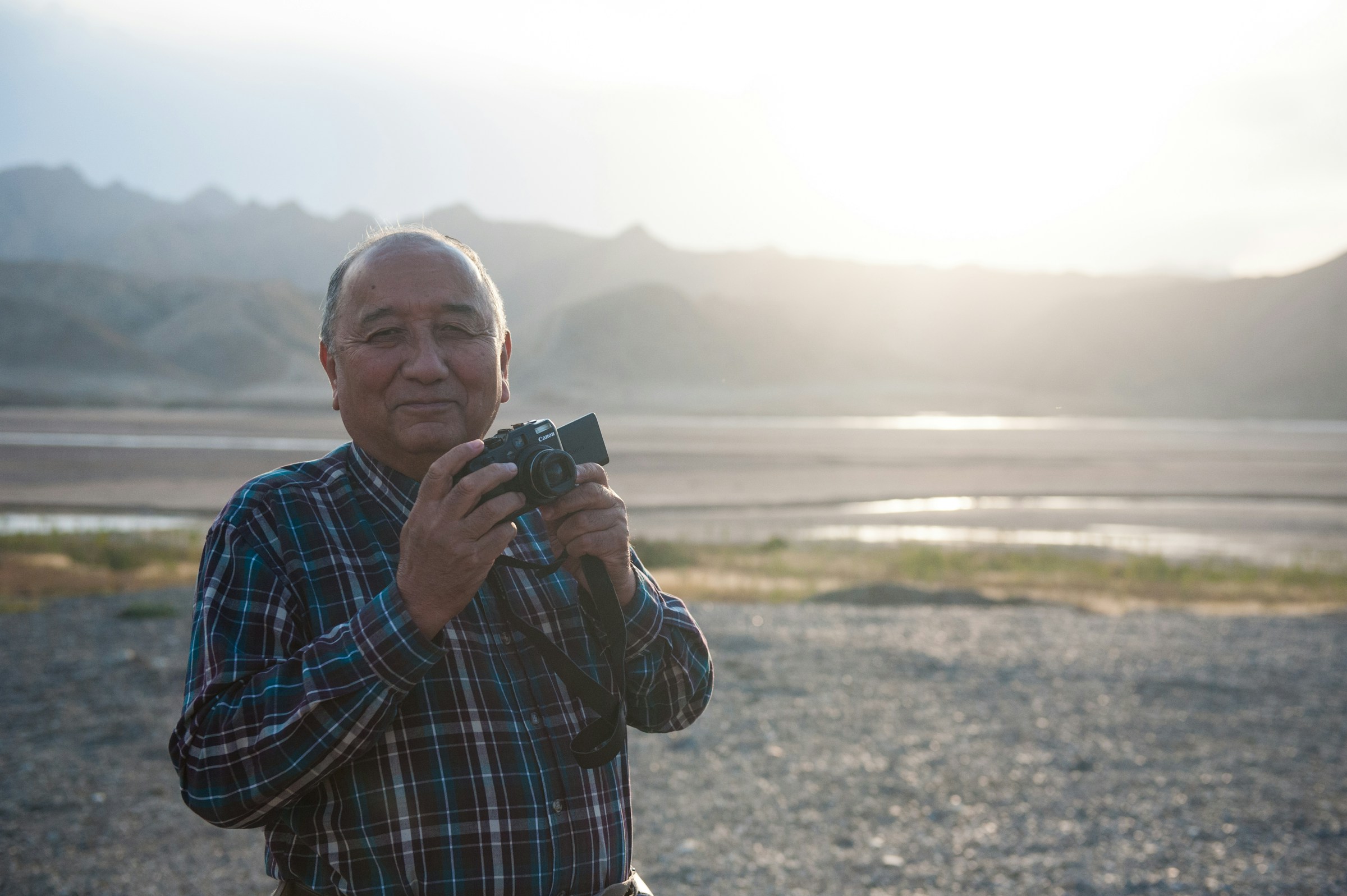An elderly man stands on the shore of a lake during sunrise, holding a camera.