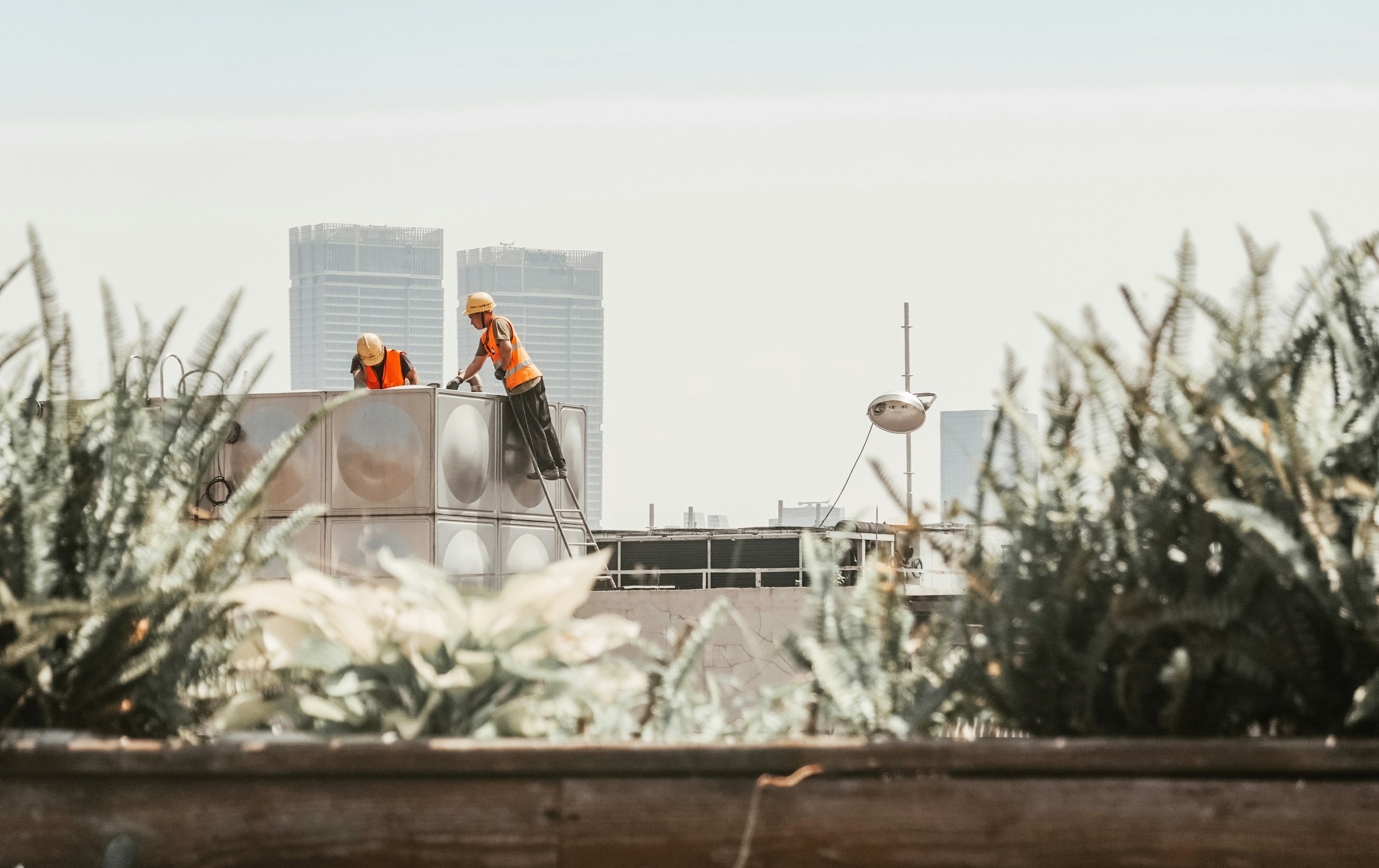 Two construction workers are seen working on the top of a building. Two city skyscrapers are seen in the background.