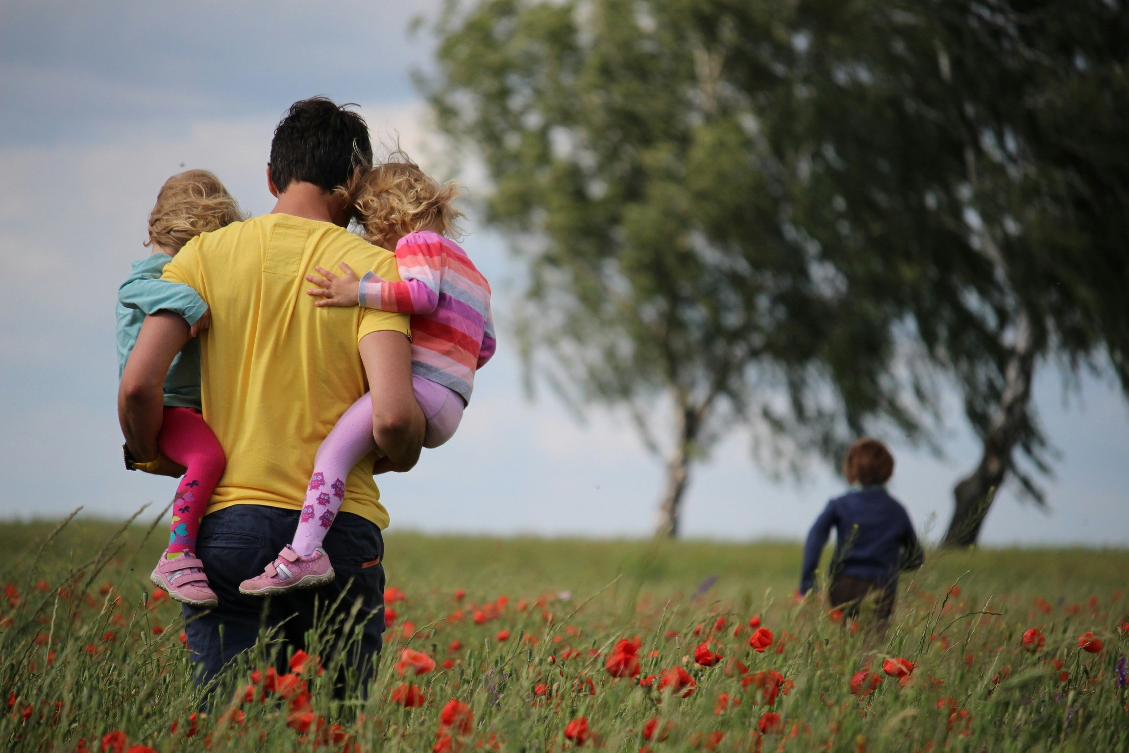 A dad is seen carrying two of his kids and walking through a field. His third kid is seen running in front of him.