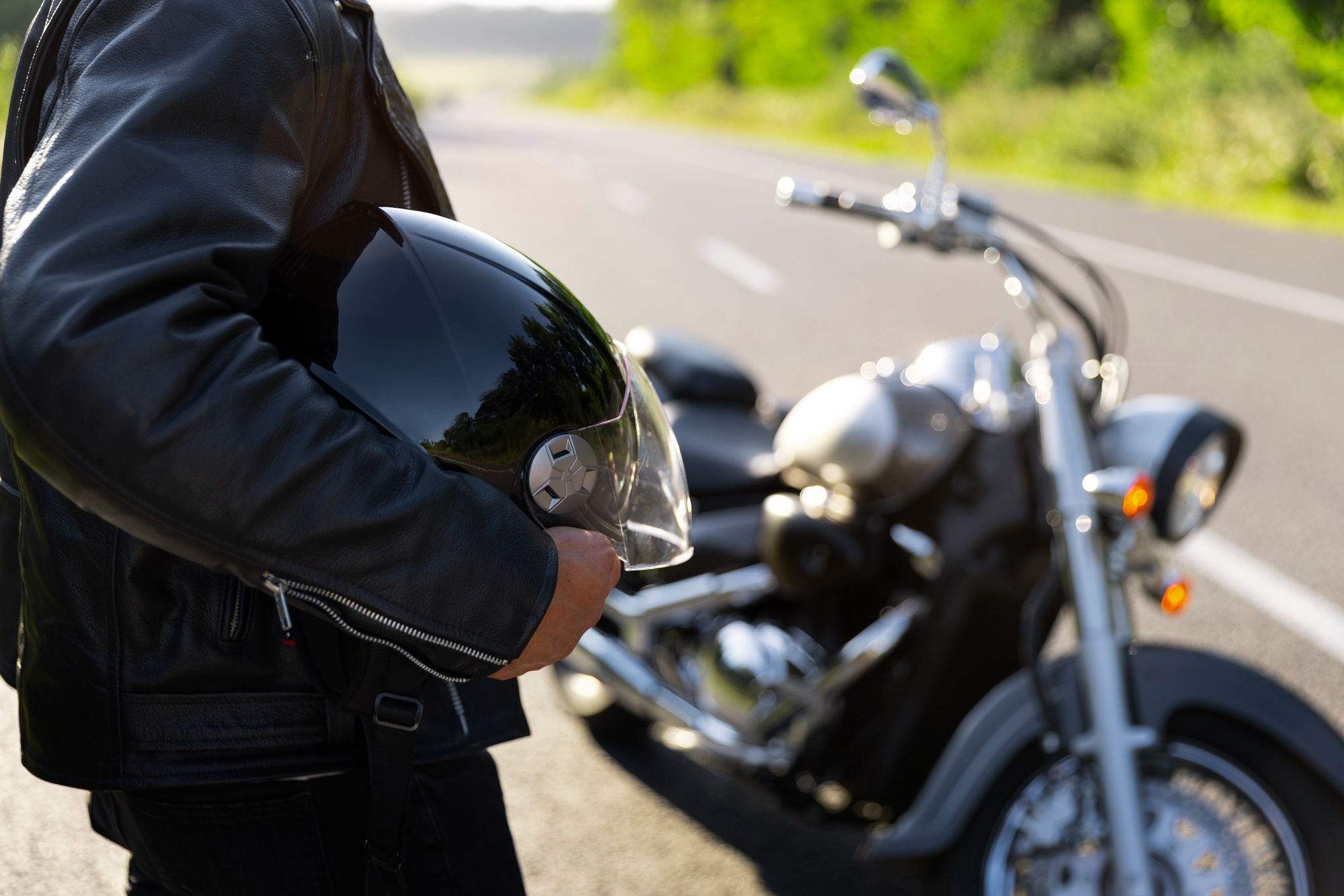 A motorcycle rider stands next to his parked motorcycle on the side of a road. He's holding his helmet in his hands.